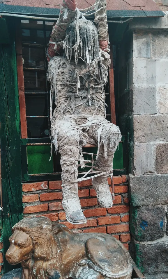 Bir kapının önünde, sargılara sarılmış bir heykel ve taş bir aslan. Bu köşeden geçen herkes bir kez durup bakıyor.