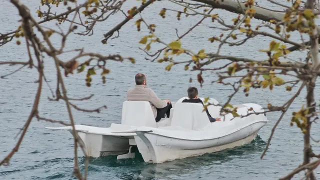 Dalların arasından, gölün ortasında pedallı teknede iki kişi.
