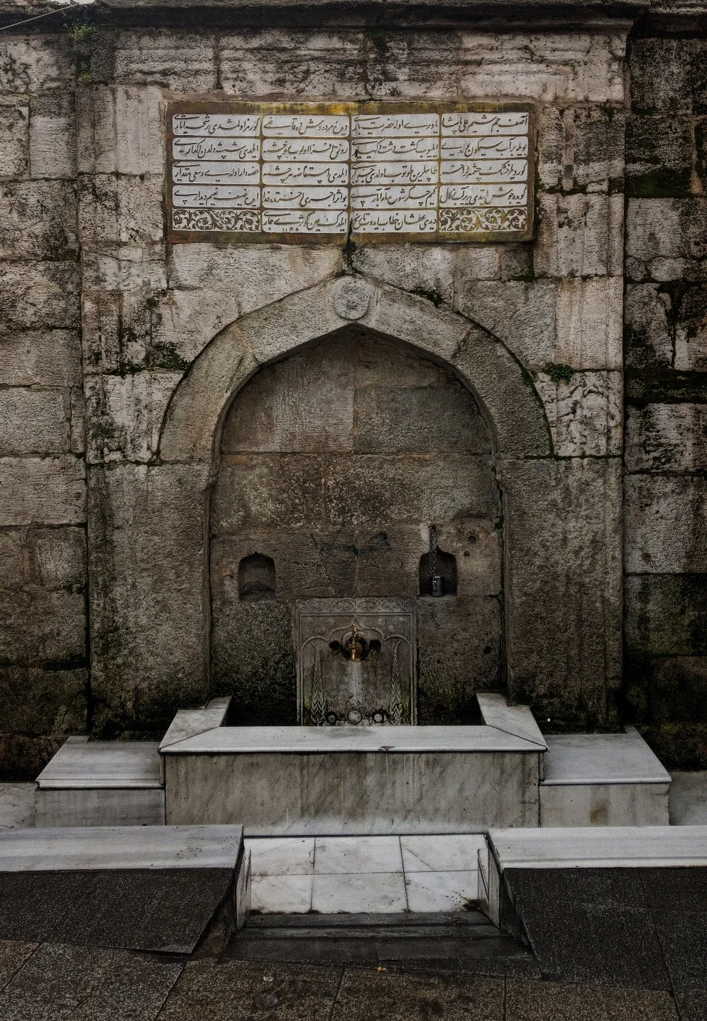 Kitabesiyle eski bir çeşme. Taşın üzerindeki her leke ayrı bir yüzyıldan kalmış gibi.